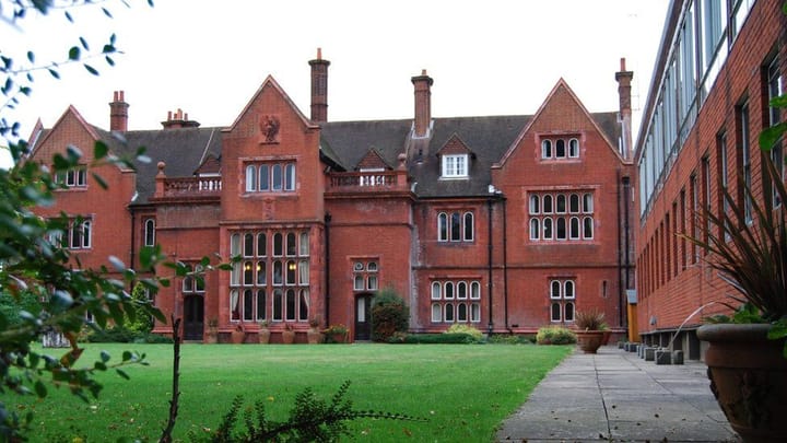 Photo of a redbrick Victorian building with a dark brown roof, in front of a green lawn with a more modern redbrick building on the right-hand edge of the photo.