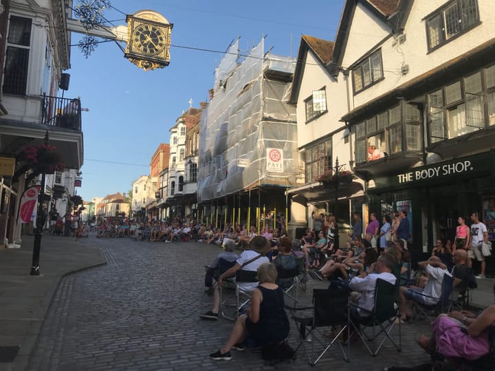 Photo of an audience sat in a semicircle on a cobbled street facing the balcony of the medieval Guildhall on Guildford High Street.